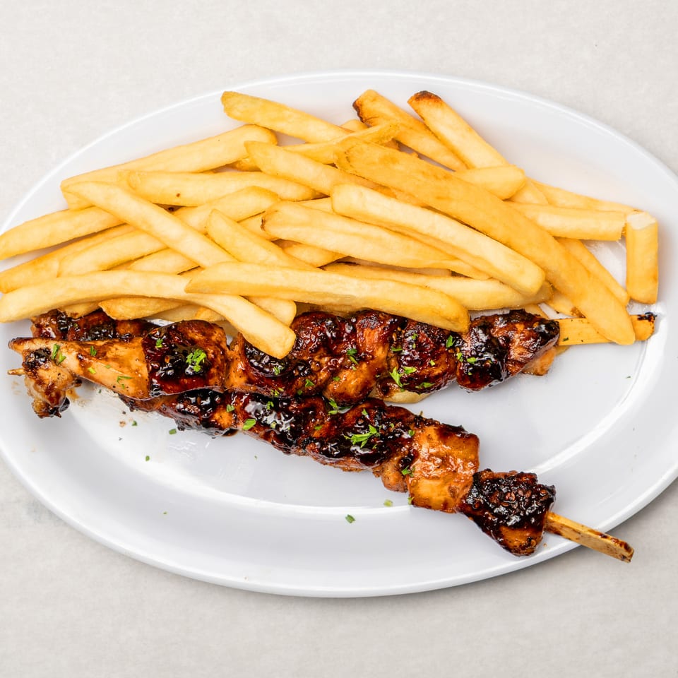 Glazed grilled chicken skewers garnished with parsley and served with golden French fries on a white plate.