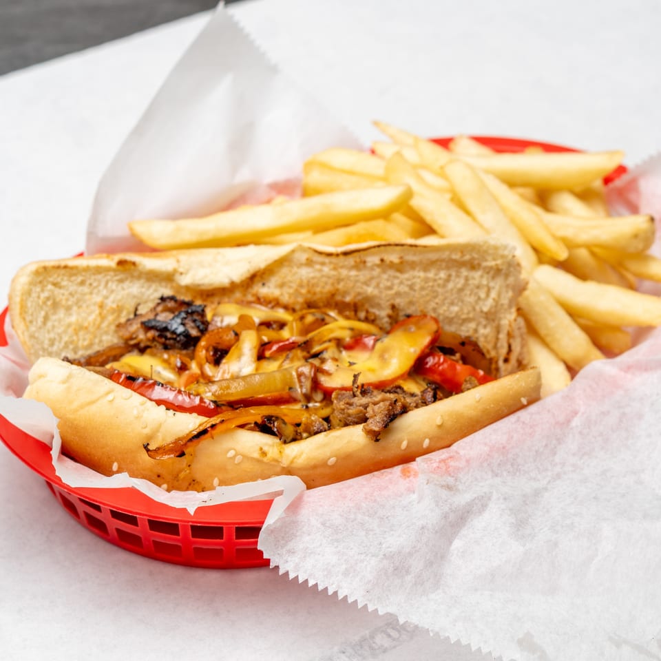 Cheesesteak sandwich with grilled peppers and onions served alongside golden fries in a red basket.