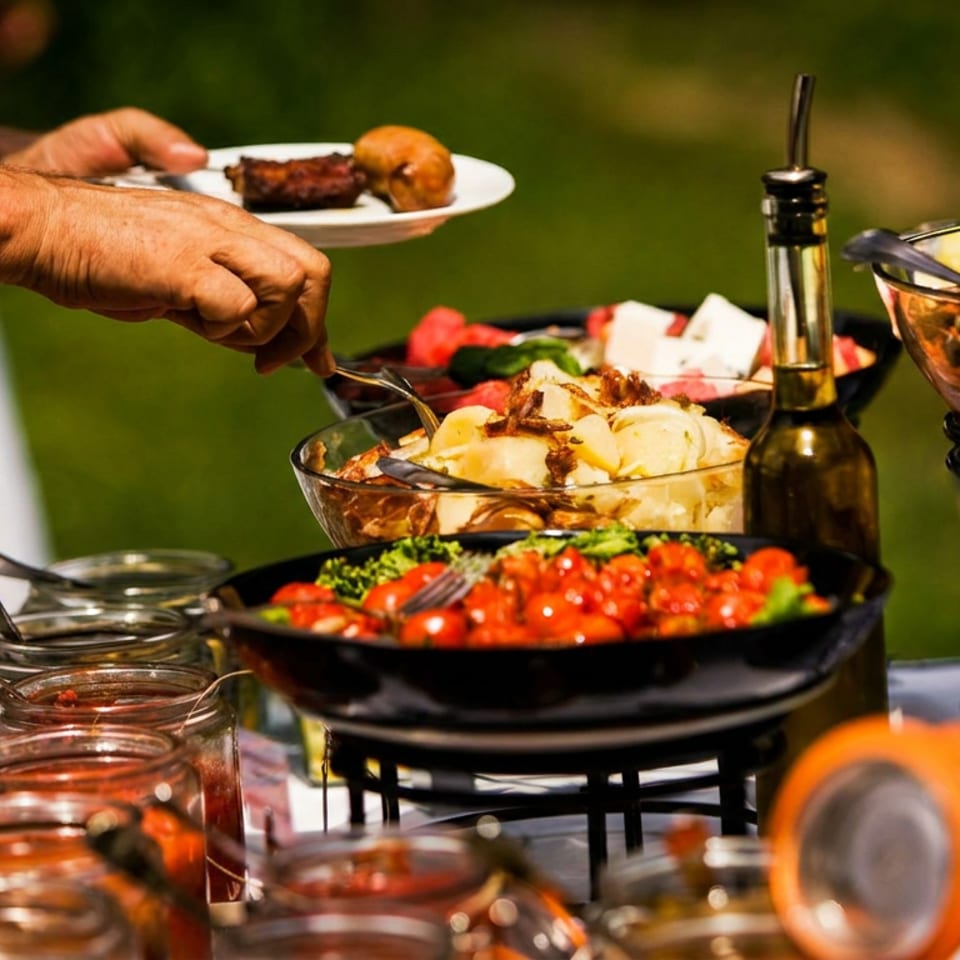 Outdoor buffet featuring salads, pasta, sauces, and grilled food, with a person serving themselves from the dishes.