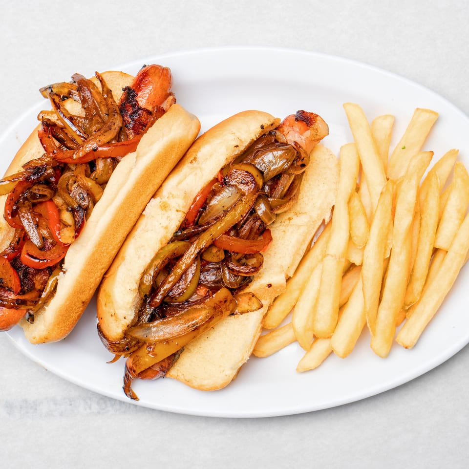 Two grilled sausages topped with sautéed peppers and onions in sandwich rolls, served with French fries on a white plate.